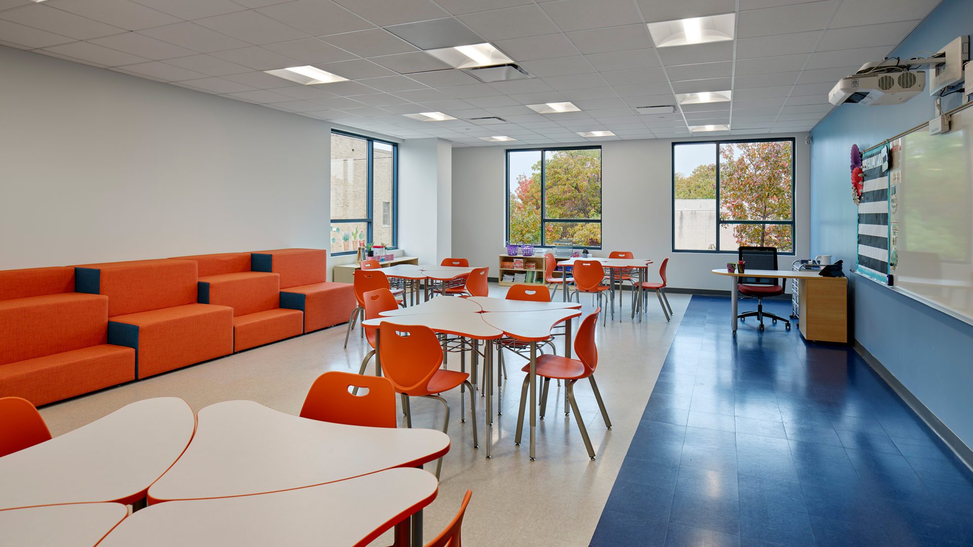 school room with white tables and orange chairs. comfy orange seats line the wall and the windows are big and bright