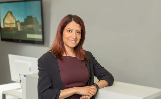 Kiran Sharma-Boon leans against a desk and smiles at camera. Wearing black blazer and burgundy blouse