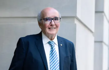 A man with glasses, a white shirt, blue jacket and pocket kerchief in front of a white background.