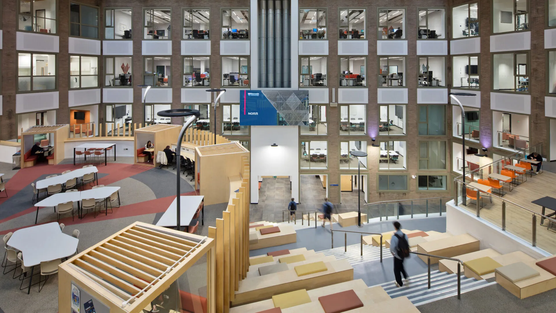 Interior of the Stephenson Building, featuring a central staircase with wooden seating and colorful cushions. Surrounding the staircase are study pods, open desks, and upper levels with glass-walled offices and classrooms.