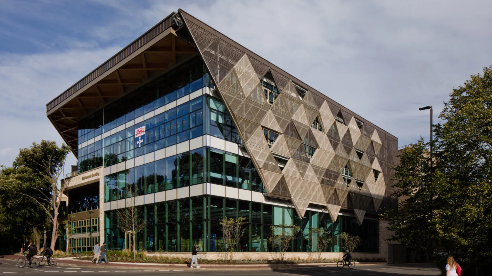 The exterior of the Stephenson Building, showcasing its modern design with a triangular metal facade, large glass windows, and a sloped roof. People are walking and cycling along the street in front of the building.