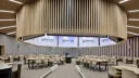 A circular lecture room within the building, featuring a suspended, slatted wooden ceiling and multiple screens displaying “Newcastle University.” Tables with chairs are arranged for collaborative learning.