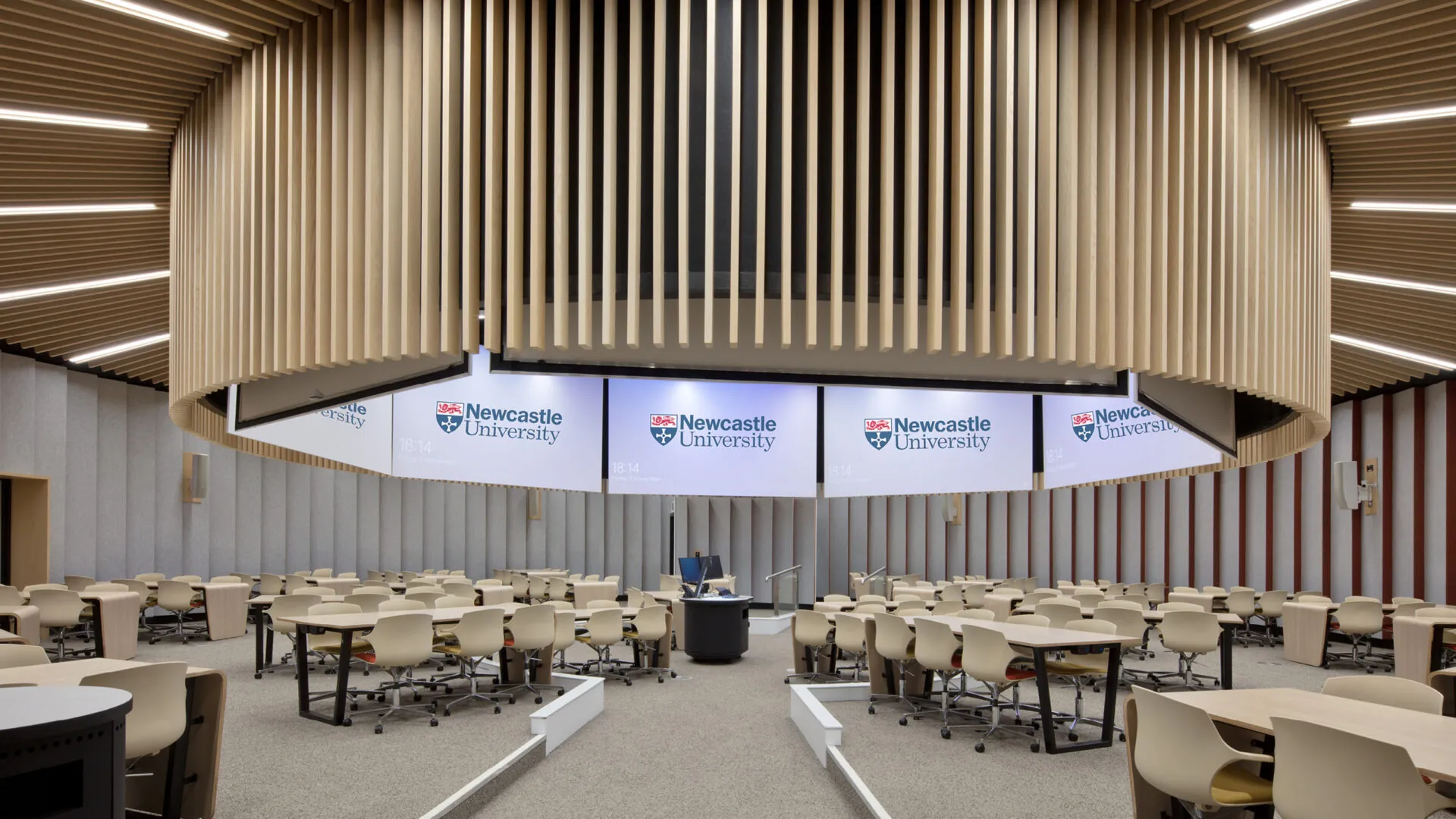 A circular lecture room within the building, featuring a suspended, slatted wooden ceiling and multiple screens displaying “Newcastle University.” Tables with chairs are arranged for collaborative learning.