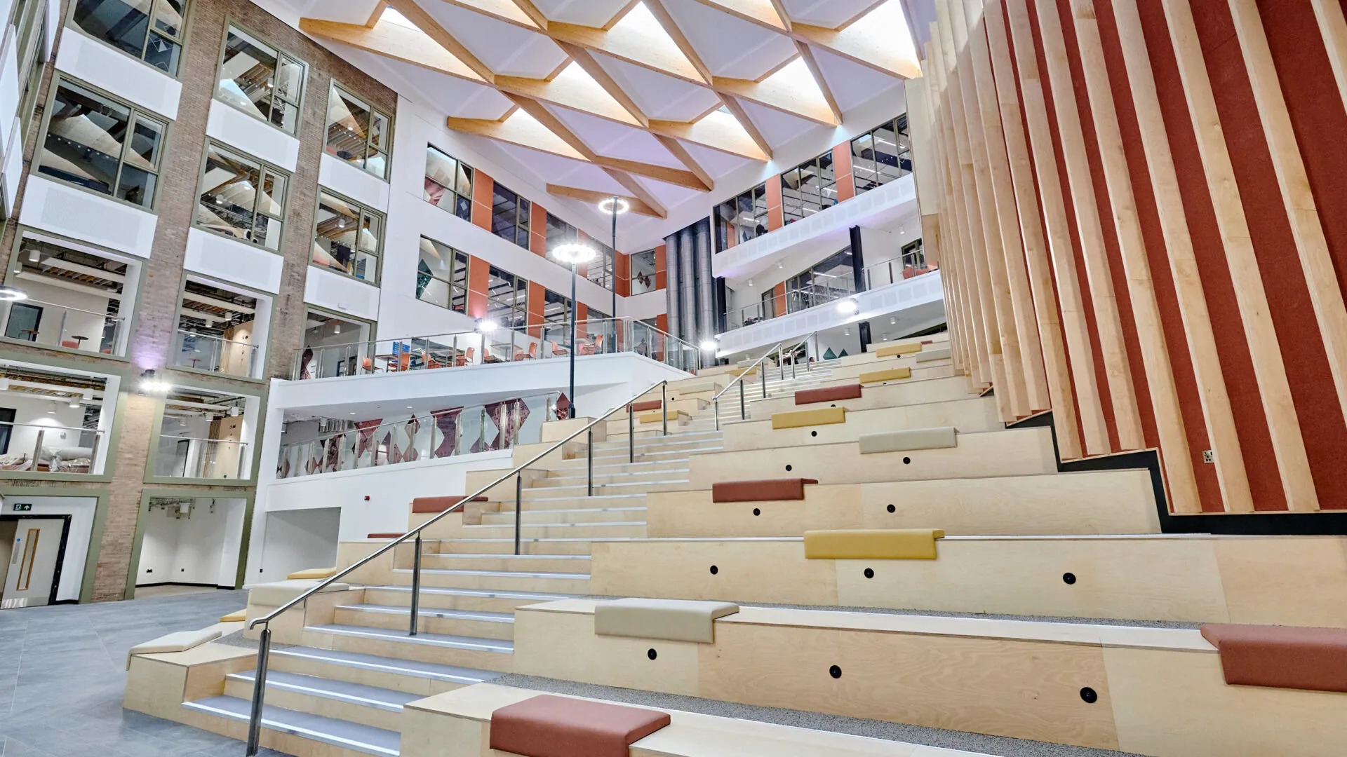 A modern atrium with a wooden ceiling and geometric beams. The central staircase and seating area are illuminated by natural light, with open spaces for socializing and studying.