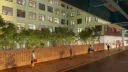 A view of a courtyard surrounded by a green building. The space includes brick walls, string lights, and a sidewalk under an overpass, with people walking and enjoying the urban setting.