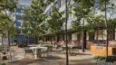 An outdoor patio area featuring tables and chairs surrounded by lush greenery. A barbecue station and trees provide a relaxing, inviting atmosphere.