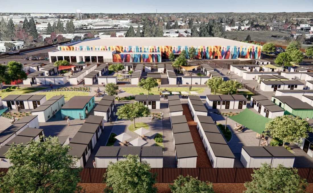 An aerial view of a community of small modular homes arranged in a grid pattern, with a colorful mural on a large building in the background. The area includes green spaces, trees, and people walking around.