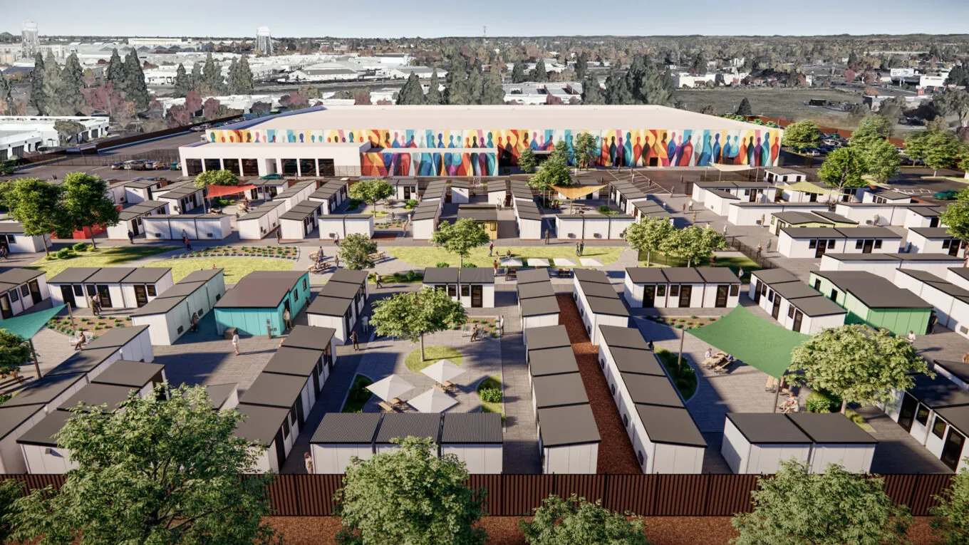 An aerial view of a community of small modular homes arranged in a grid pattern, with a colorful mural on a large building in the background. The area includes green spaces, trees, and people walking around.