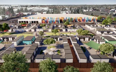 An aerial view of a community of small modular homes arranged in a grid pattern, with a colorful mural on a large building in the background. The area includes green spaces, trees, and people walking around.