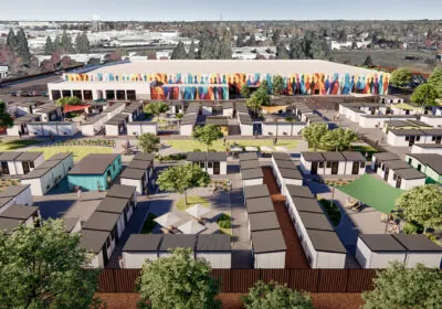 An aerial view of a community of small modular homes arranged in a grid pattern, with a colorful mural on a large building in the background. The area includes green spaces, trees, and people walking around.